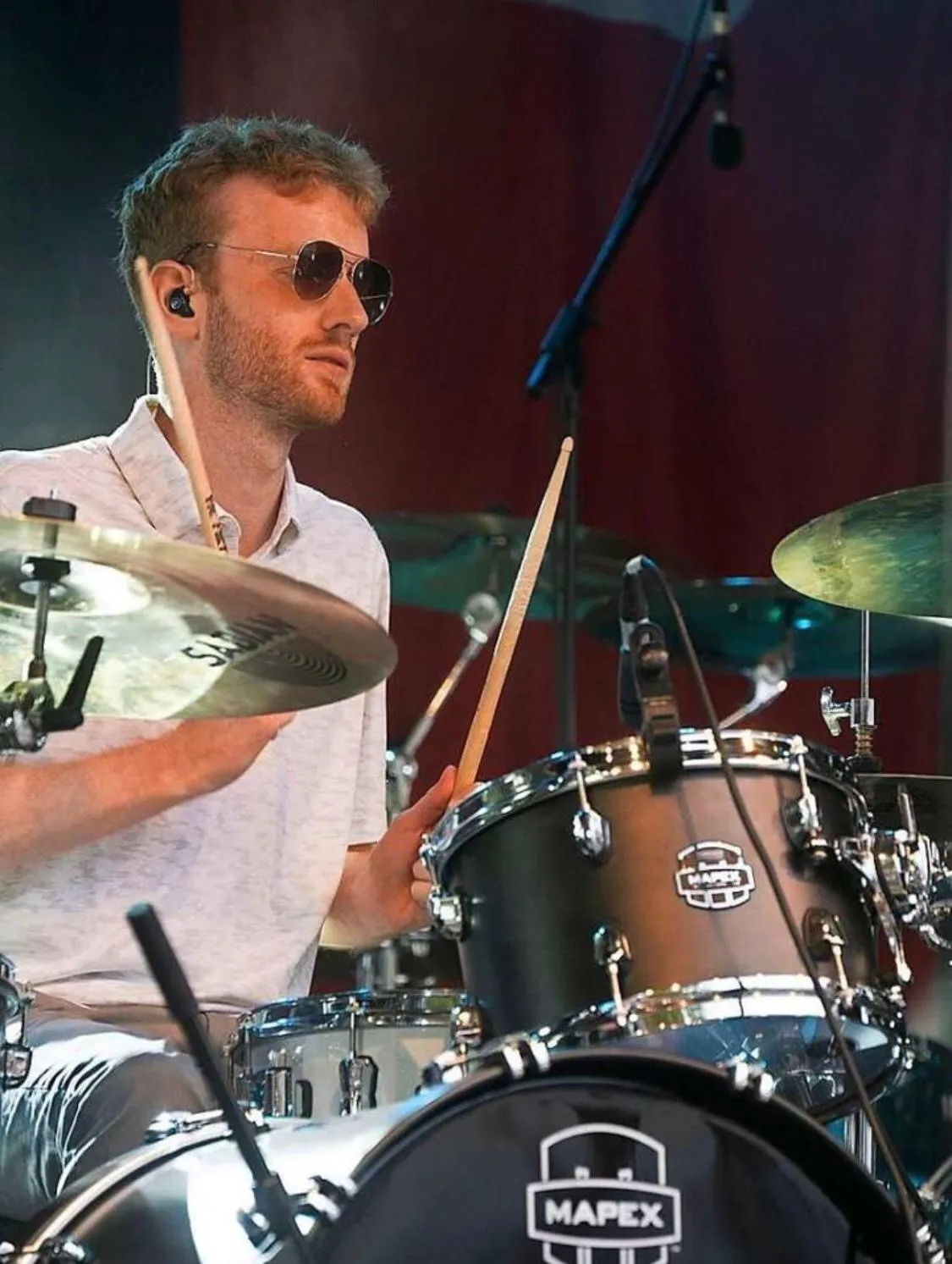 Man wearing sunglasses playing a Mapex drum set with drumsticks in a live music setting.