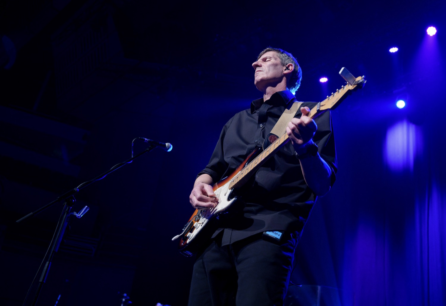 Male musician playing electric guitar on stage under blue and purple lights.