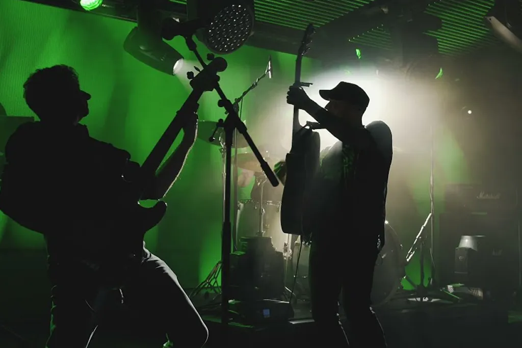 Silhouettes of two musicians playing guitars on stage with green and white backlighting and a drum set in the background.